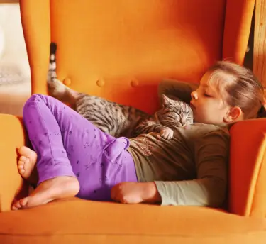 Little girl sleeping with a tabby cat on an orange armchair, suggesting cat fur as an allergy trigger